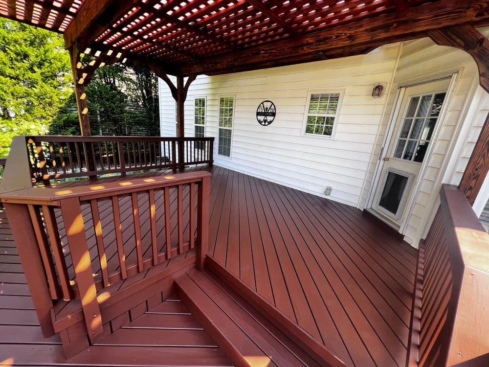 Spacious wooden deck with a pergola and white house exterior, featuring sunlight and greenery.