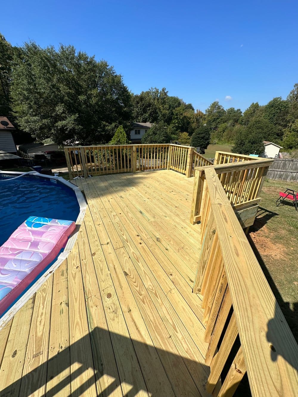 Wooden deck overlooking a backyard pool with blue sky and greenery in the background.