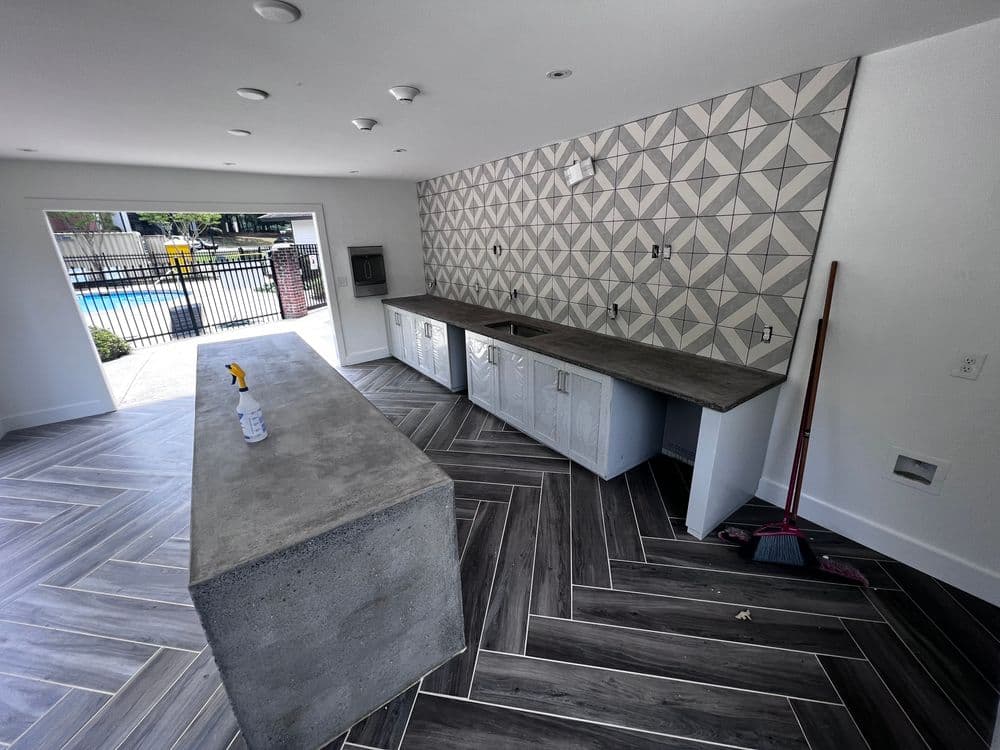 Modern kitchen interior with geometric tiled wall, concrete island, and wood-like flooring.