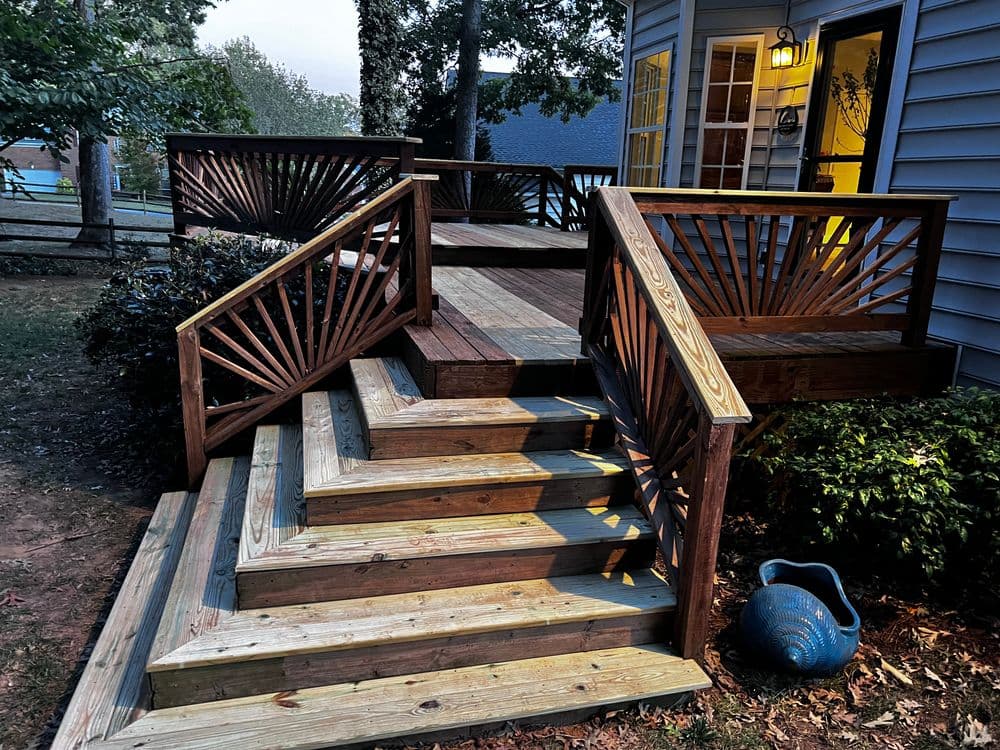 Wooden deck stairs with decorative railings, illuminated by outdoor lighting, surrounded by greenery.