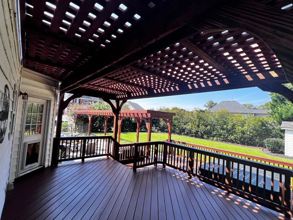 Spacious wooden deck with pergola, surrounded by greenery and clear blue skies.