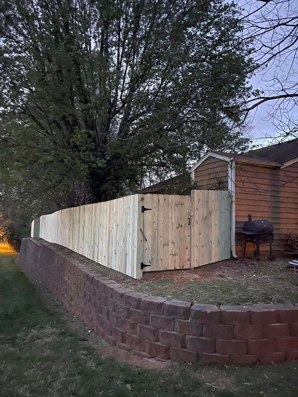 New wooden fence with black gate, set against a brick wall and tree at dusk.